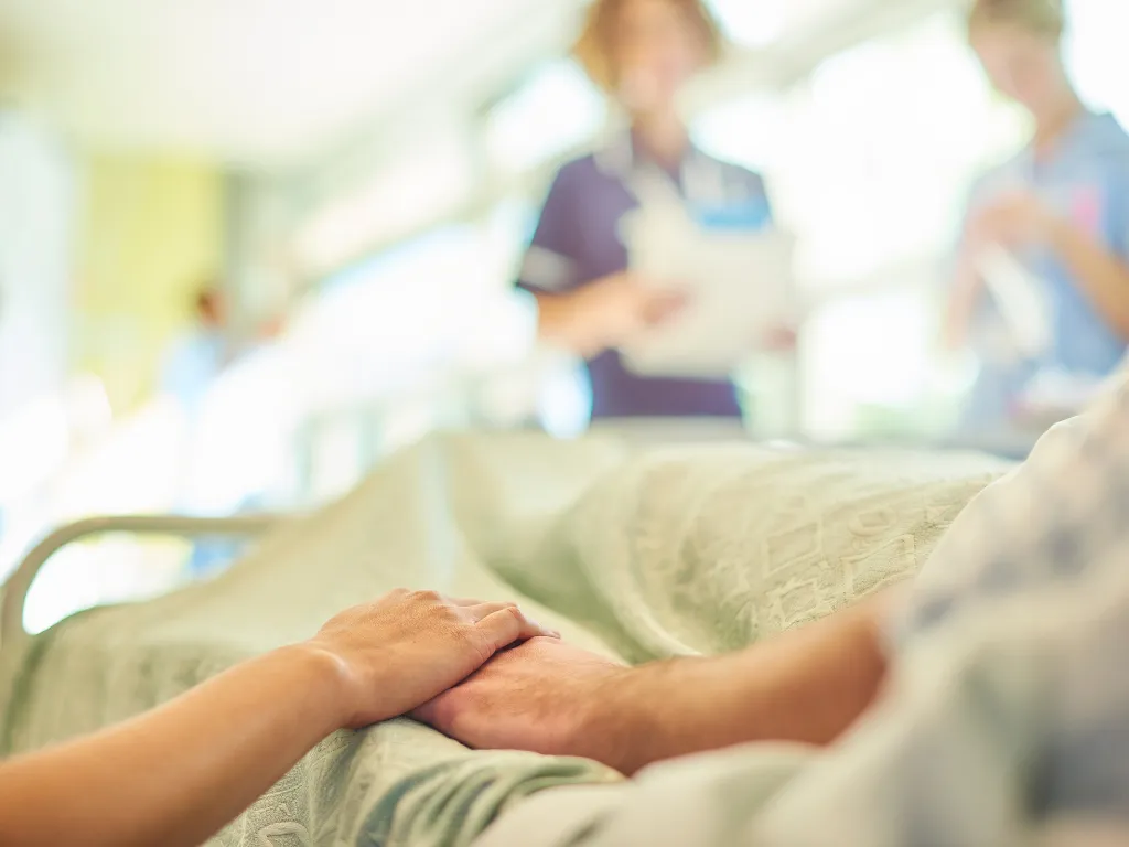 Close-up of a patient lying in a hospital bed holding hands with a loved one, while healthcare staff stand in the background.